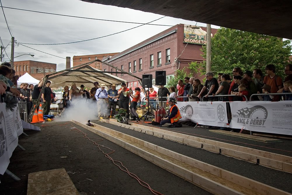 A group of people gathered under a bridge for an event, with tents and banners visible. The setting is urban with brick buildings in the background.
