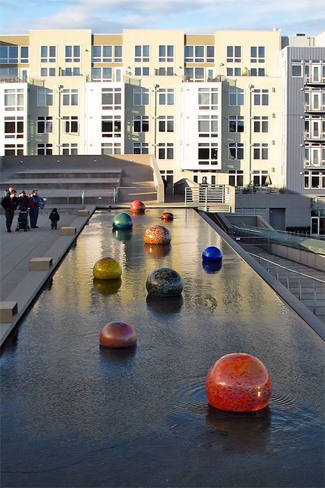A serene urban water feature reflecting a modern building, adorned with vibrant, colorful spheres. People stroll along the pathway, enjoying the tranquil setting under a clear sky.