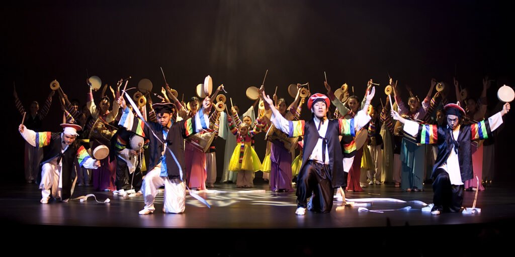 A group of performers dressed in vibrant traditional attire, engaging in a dynamic dance routine on a dimly lit stage.