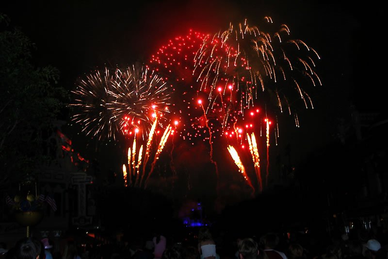 Bright red and white fireworks explode vibrantly against a dark night sky, illuminating the scene. Onlookers in the foreground watch from the shadows below.