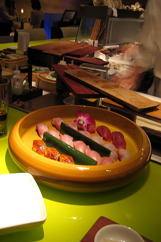 A colorful sushi platter featuring an assortment of nigiri and sashimi on a vibrant green countertop. The scene depicts a lively sushi restaurant atmosphere with a chef preparing food in the background.