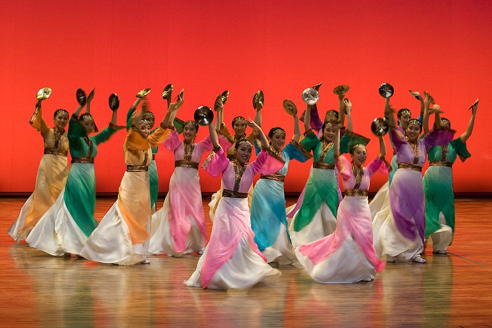 A group of dancers in vibrant, flowing outfits performing a synchronized routine on a wooden stage against a red backdrop. Each dancer holds props above their heads as part of the choreography.
