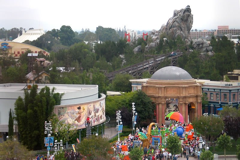 A view of a lively amusement park with colorful rides and a rocky mountain structure. The scene includes people walking around and enjoying the attractions.
