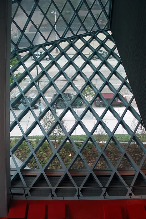 A geometric lattice structure viewed from inside, with a street scene visible through the pattern. Red and grey elements frame the shot.