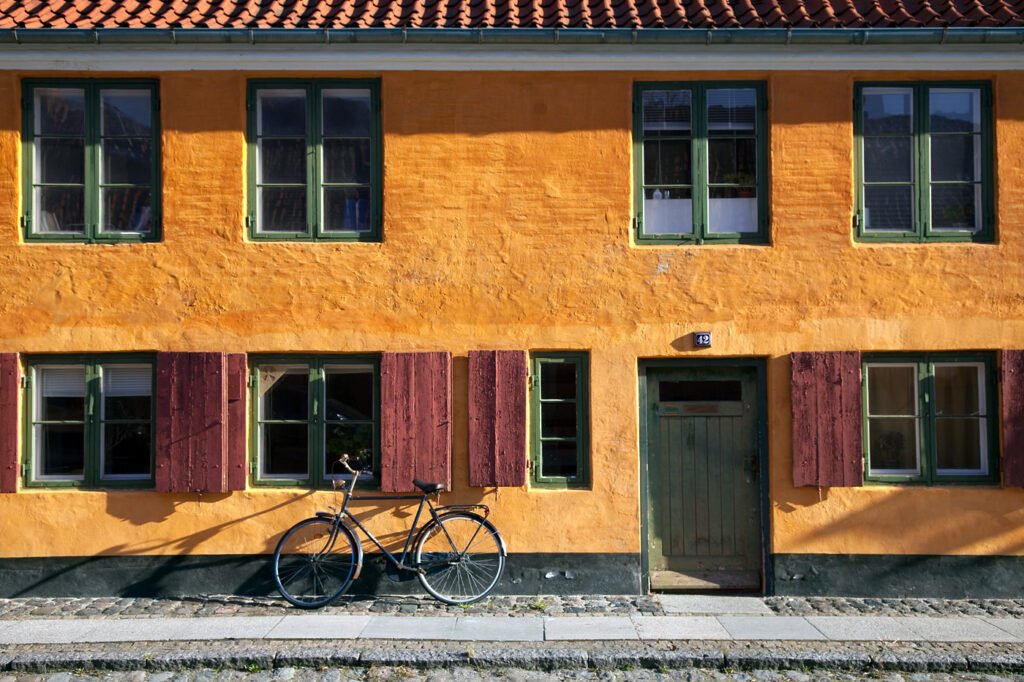 A classic bicycle leans against a vibrant orange building, adding timeless charm. Green shutters frame the windows.
