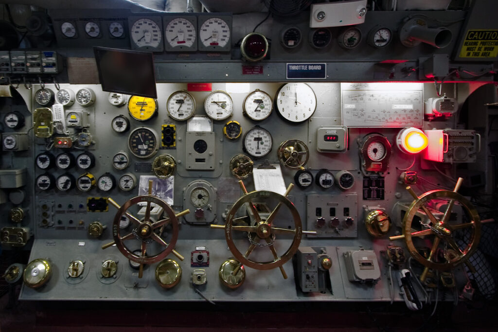 A detailed view of an old ship's control panel featuring brass steering wheels, numerous gauges, and illuminated indicators. The panel is set in a dark environment with a warm red glow.