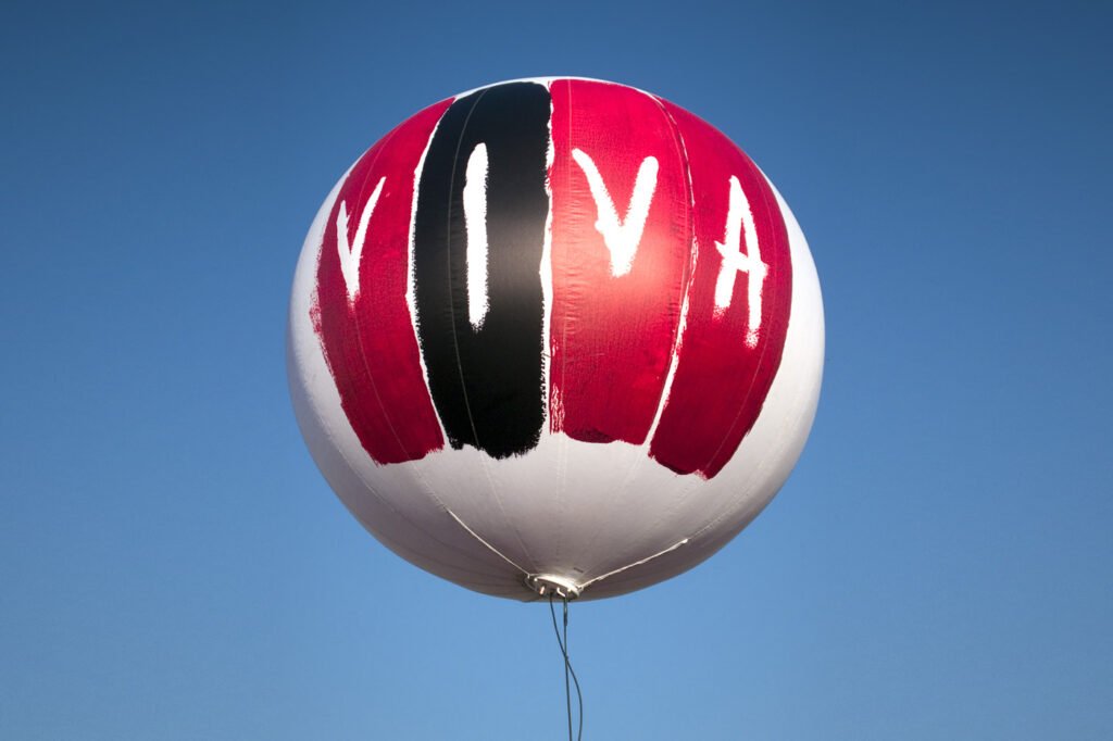 A red, white, and black balloon with the word 'Viva' floats against a clear blue sky.