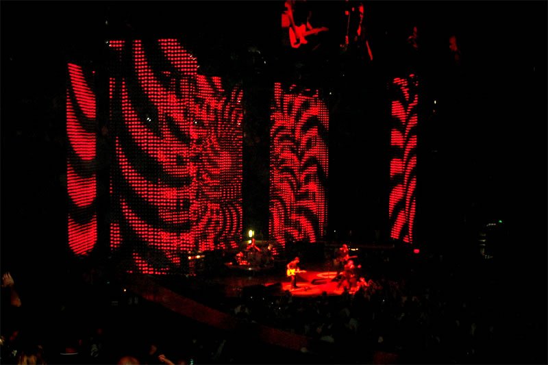 A nighttime outdoor scene featuring a striking light display with red patterns forming large letters or decorative elements against a dark background.
