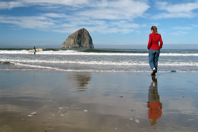 A person in a red jacket walks along the wet sand toward the iconic Cannon Beach sea stack. Their reflection is mirrored on the smooth tidal surface beneath them.