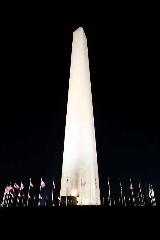 The Washington Monument stands illuminated against a dark sky with flags around its base at night.
