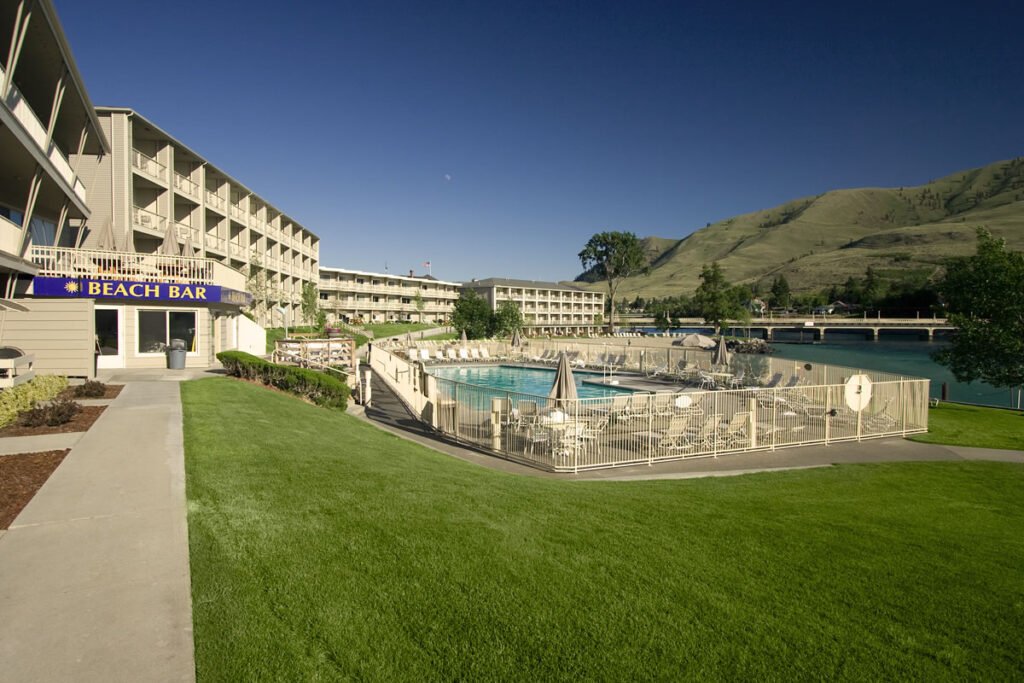 An outdoor pool area next to a waterfront hotel, surrounded by green lawns and hills.