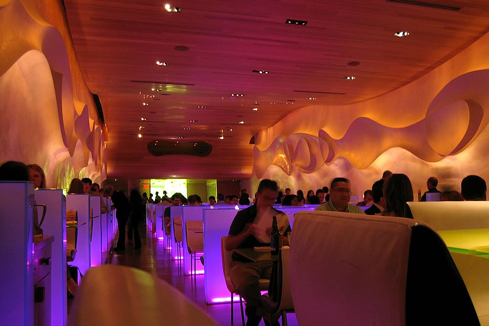 A uniquely designed indoor space featuring wavy, illuminated ceiling and ambient lighting. People are seated at tables, enjoying the atmosphere in what appears to be a restaurant or event venue.