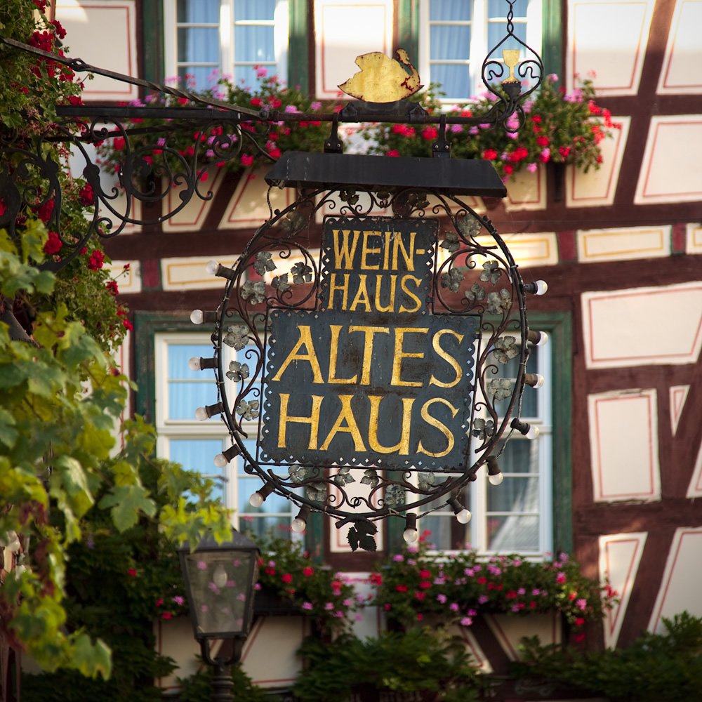 A circular sign reading 'Weinhaus Altes Haus' hangs in front of a half-timbered building with red flowers.