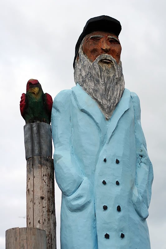 A quirky sculpture of a bearded fisherman in a blue coat with a parrot perched on a wooden post beside him.