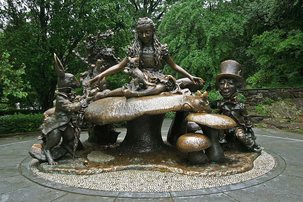 A charming bronze garden statue featuring characters sitting around a mushroom table, set in a lush green garden. This detailed sculpture depicts fairy tale-like figures enjoying a whimsical gathering.
