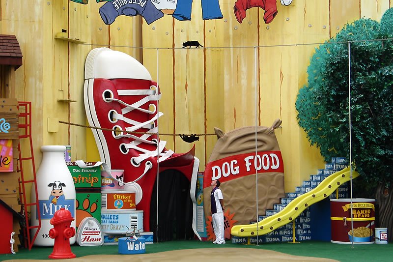 A playful outdoor scene featuring a giant red shoe, a large dog food sack, and a yellow slide. A person stands near the slide, surrounded by oversized objects like a milk bottle and a fire hydrant.