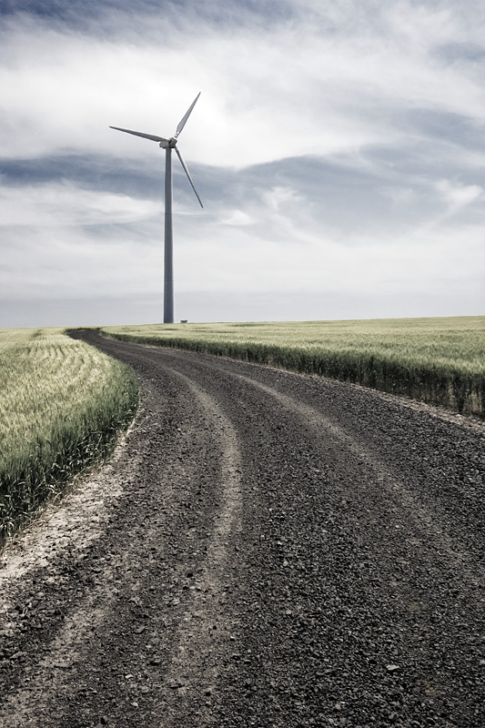 An image capturing a gravel road extending into the distance with a solitary wind turbine standing prominently in the middle of a flat, expansive landscape under a cloudy sky.