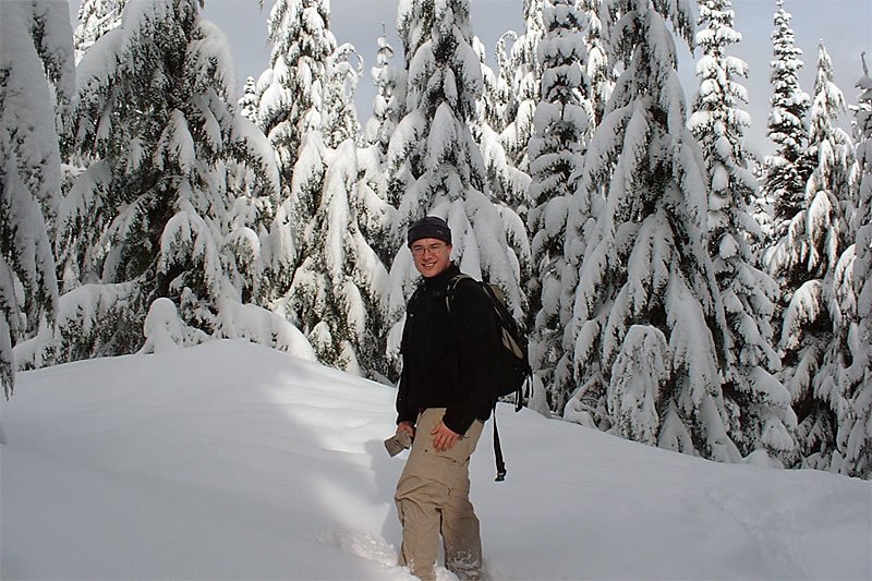 A hiker stands amidst snow-covered trees in a serene winter landscape. The ground is blanketed in fresh snow, creating a peaceful atmosphere.