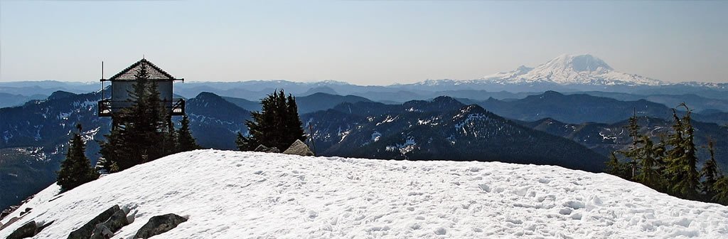 A picturesque snowy mountain landscape featuring a fire lookout tower and distant rolling hills under a clear blue sky.