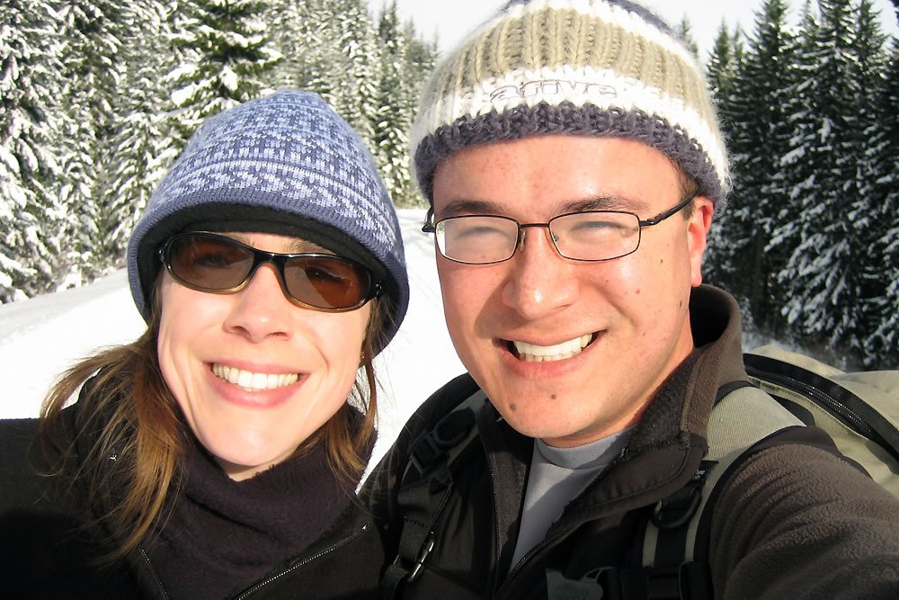 A couple bundled up in winter gear smiles at the camera amidst a snowy forest backdrop. They appear to be enjoying a day of outdoor activities in a picturesque winter setting.
