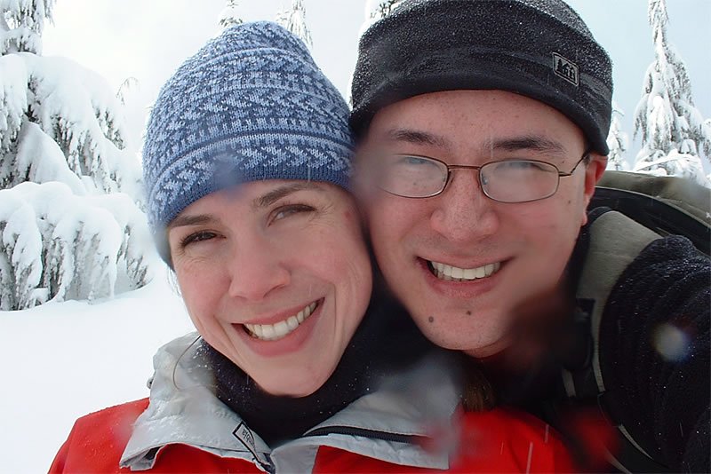 A joyful selfie of John and Kristie bundled up in winter gear, standing amidst a snowy landscape.