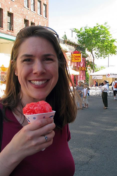 A woman smiles while holding a cup of red Italian ice. She is standing on a street with market stalls and trees in the background.