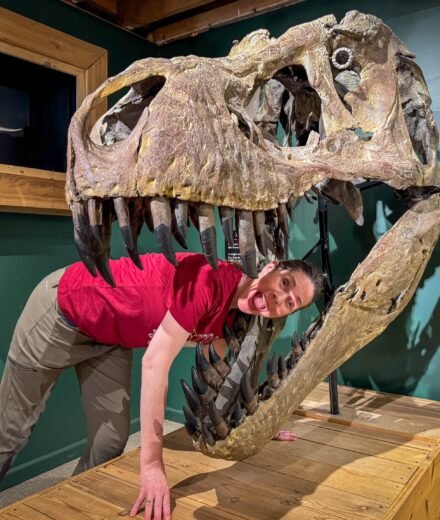 A woman in a red shirt and beige pants leans playfully against a large T-Rex skull exhibit inside a museum. The background features green walls with shadowed dinosaur silhouettes and a mounted velociraptor model behind glass.