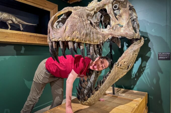 A woman in a red shirt and beige pants leans playfully against a large T-Rex skull exhibit inside a museum. The background features green walls with shadowed dinosaur silhouettes and a mounted velociraptor model behind glass.