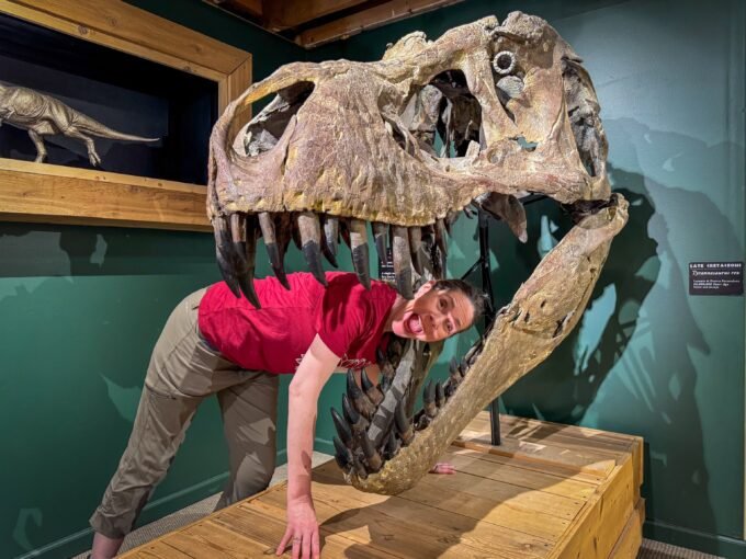 A woman in a red shirt and beige pants leans playfully against a large T-Rex skull exhibit inside a museum. The background features green walls with shadowed dinosaur silhouettes and a mounted velociraptor model behind glass.