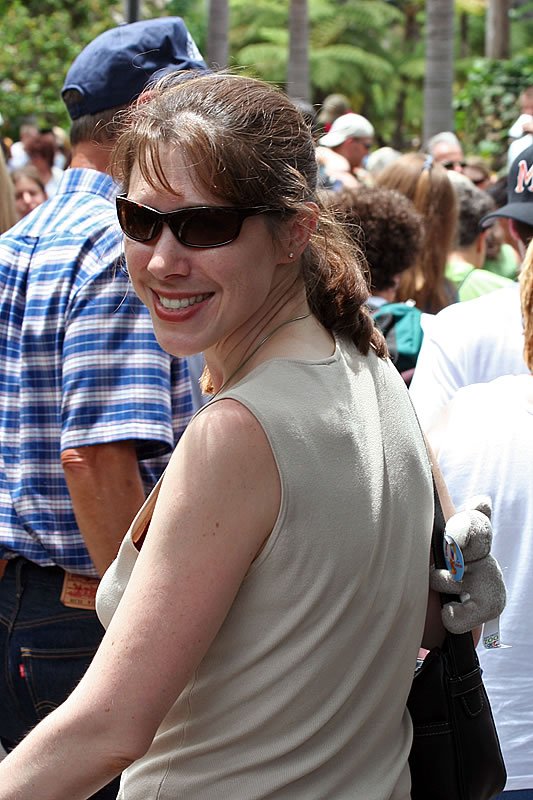 A smiling woman wearing sunglasses and a casual outfit. She appears to be at an outdoor event with a crowd of people in the background.