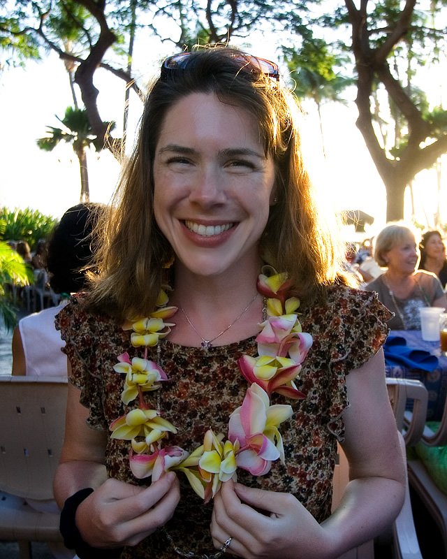 A woman smiles while wearing a colorful plumeria lei. The setting features lush trees and casual seating outdoors.