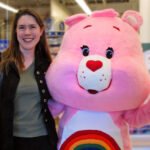 A woman stands smiling next to a pink Care Bear mascot at an indoor event. They are captured in a cheerful moment together.
