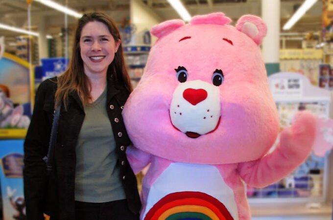 A woman stands smiling next to a pink Care Bear mascot at an indoor event. They are captured in a cheerful moment together.