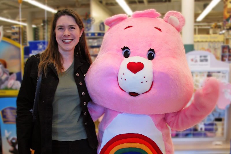 A woman stands smiling next to a pink Care Bear mascot at an indoor event. They are captured in a cheerful moment together.