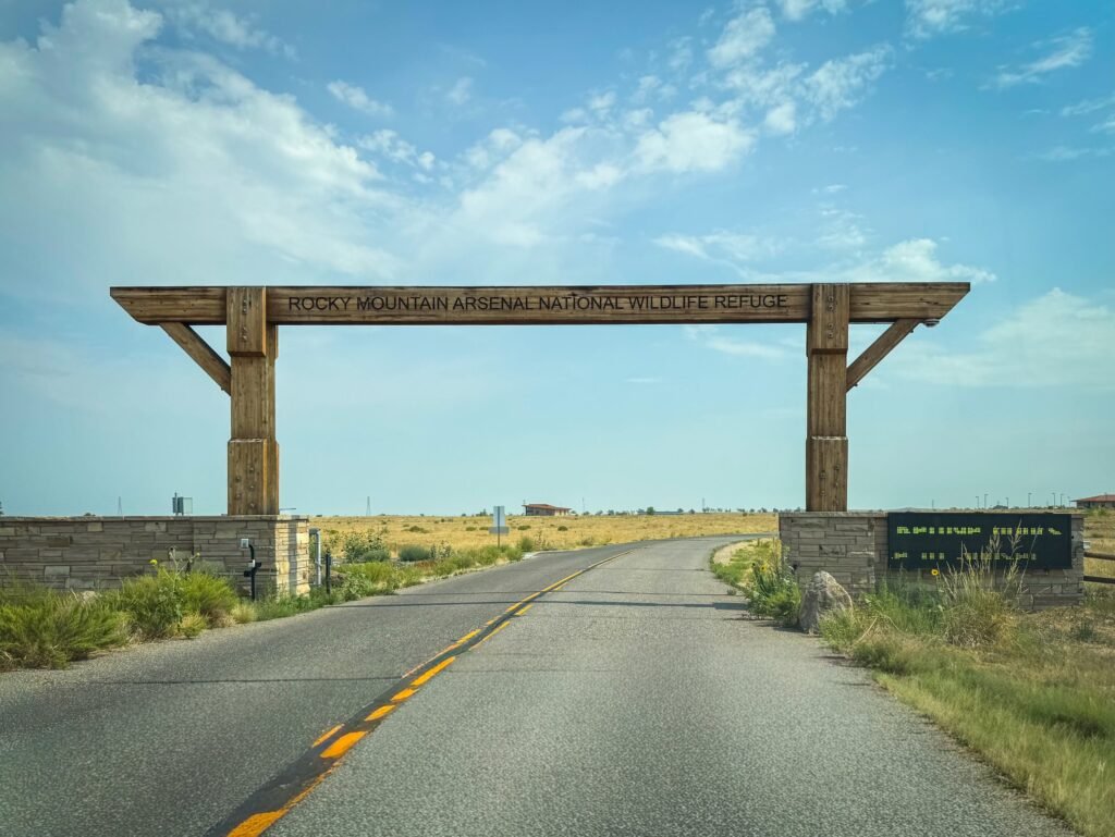 A large wooden archway marks the entrance to a scenic road flanked by grassy fields under a blue sky.