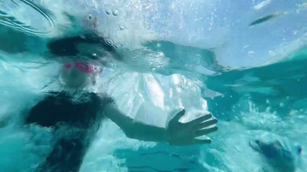 View from beneath the surface, looking up at a person swimming and splashing in clear, turquoise water.