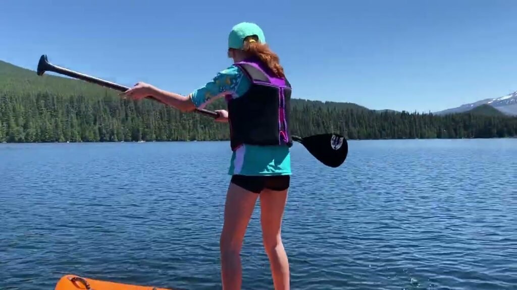 A person in a life vest stands on a paddleboard on a calm lake. The background features a clear blue sky, forested hills, and mountains.