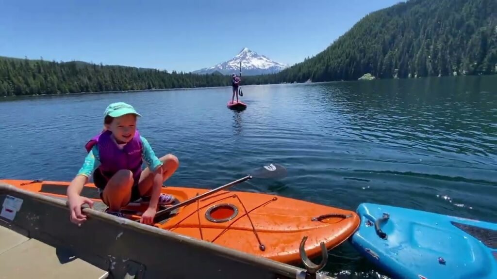 An orange and blue kayak are docked at a lake with mountains and a paddleboarder in the background, capturing a moment of water sports adventure.