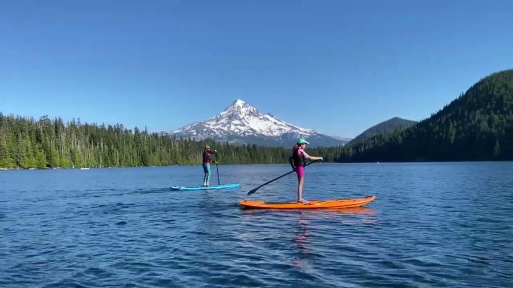 Two people stand on paddle boards on a blue lake surrounded by forested hills and a distant snow-capped mountain peak.