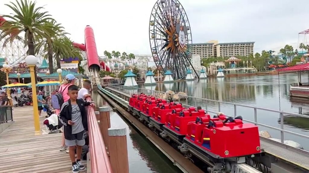 A red water slide ride with passengers enjoying the thrill near a boardwalk, with a Ferris wheel in the background.