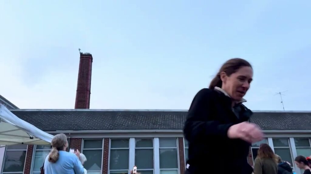 Guests socialize outside a low-rise building with a tall brick chimney under a cool blue dusk sky.