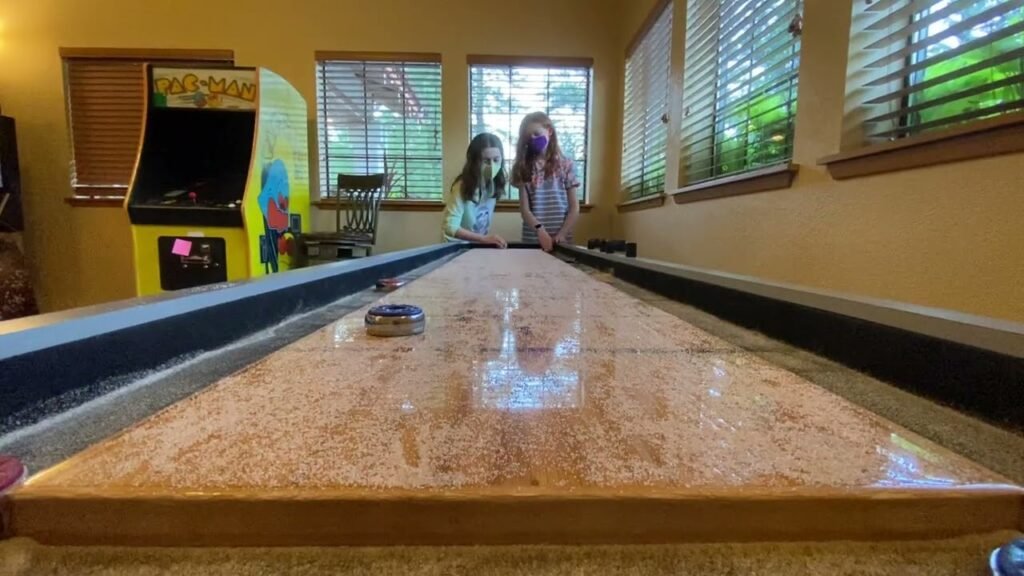 Two children playing indoor curling, with a puck on the synthetic sheet and an arcade machine nearby.
