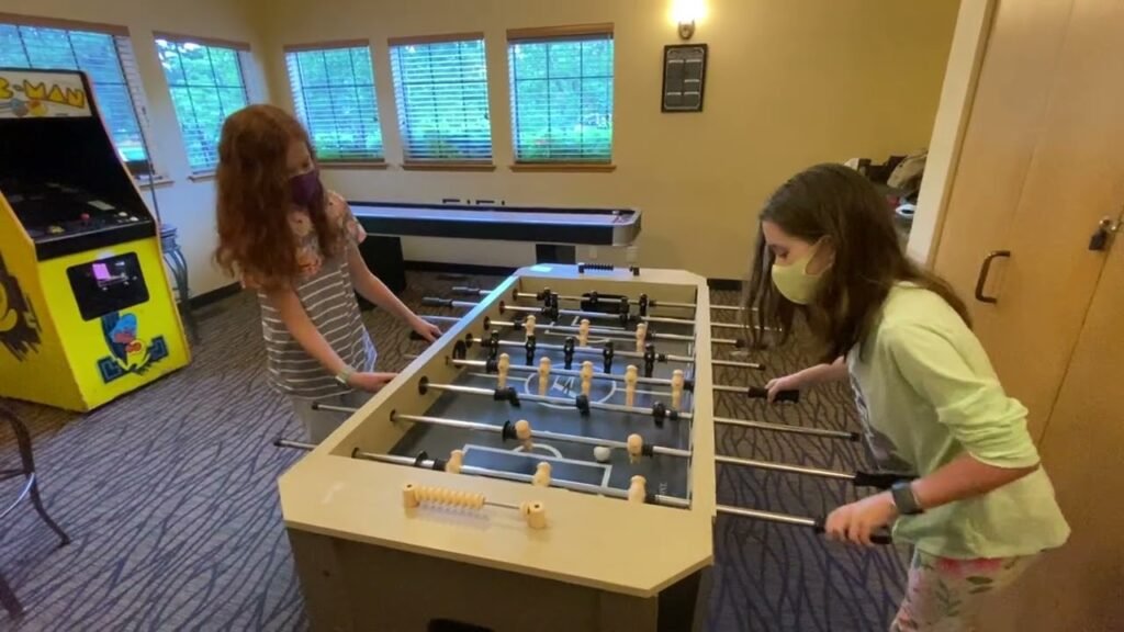 Two children playing foosball in a rec room with an arcade machine and air hockey table visible in the background.