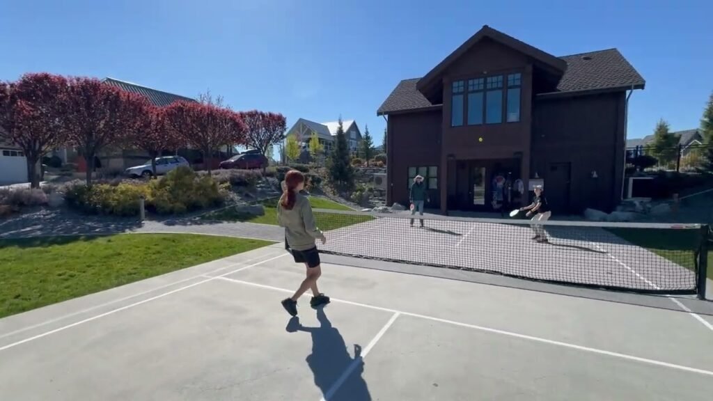 A person walking on an outdoor tennis court with a large log-style building and red-leafed trees in the background.
