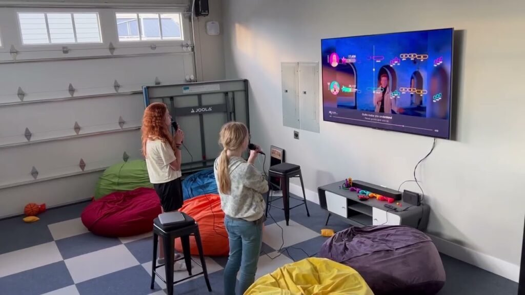 Two girls singing karaoke in a garage with bean bags, a large TV, and colorful floor tiles.