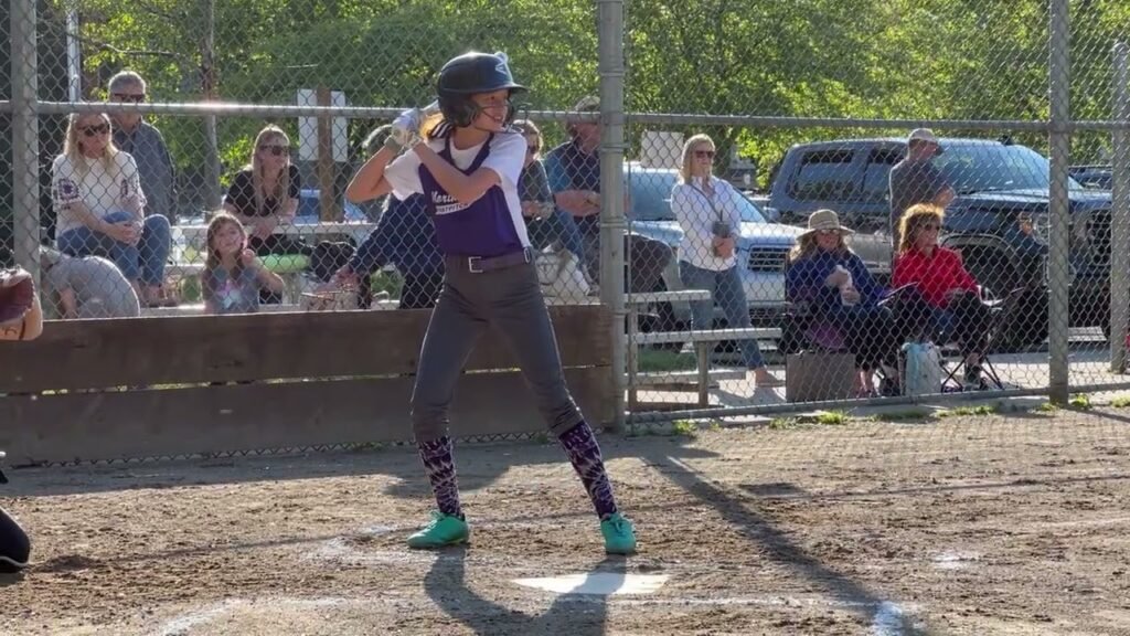 A person wearing a helmet and purple shirt prepares to bat at a local baseball field with spectators watching.