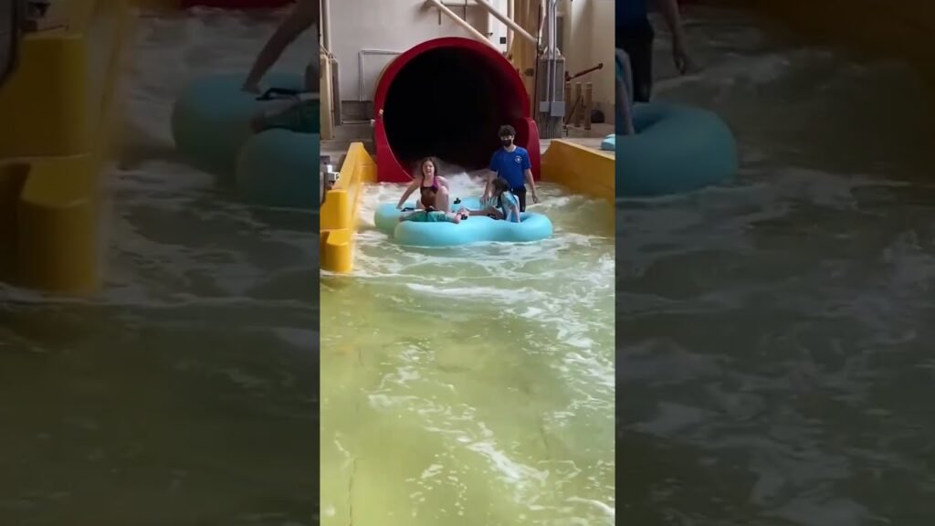Riders in a blue inner tube slide through turbulent water and exit a red tube at a water park.