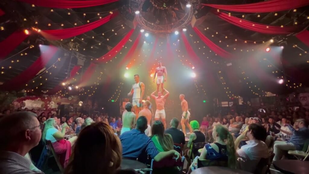 A live stage show with performers and bright red decorative drapes under colorful lights. An engaged audience watches in the foreground.
