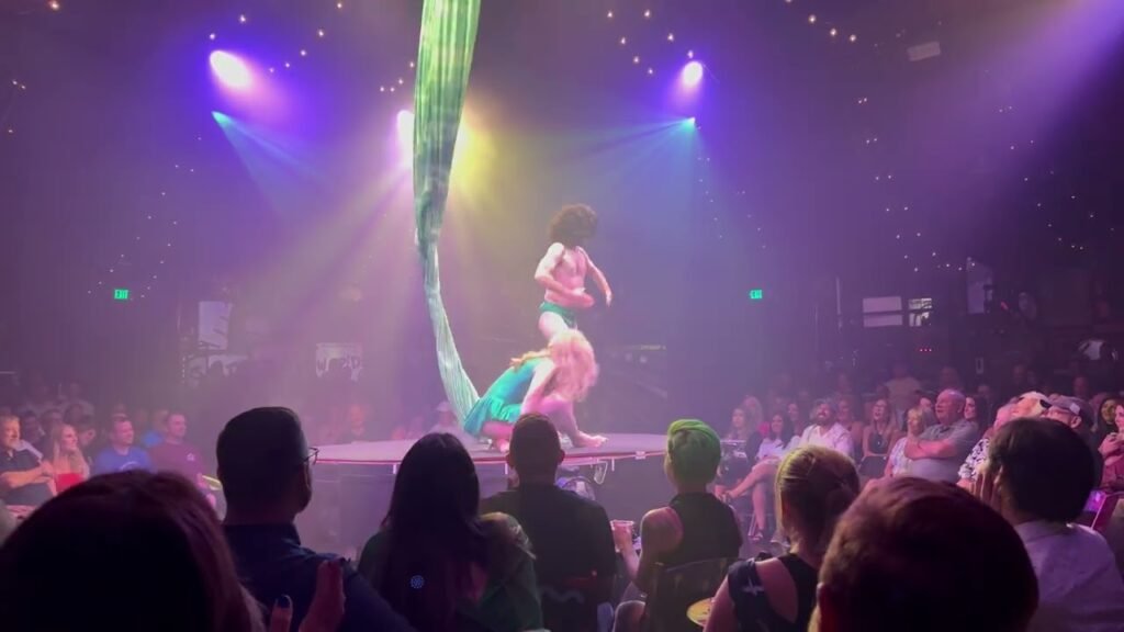 A performer in teal and pink outfit is suspended from green fabric on stage in front of cheering crowd.