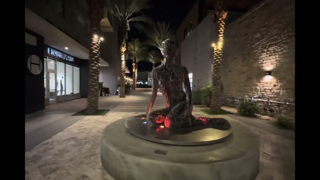A statue with a glowing base sits in a palm-lined plaza at night. Illuminated storefronts and a brick wall line the path.